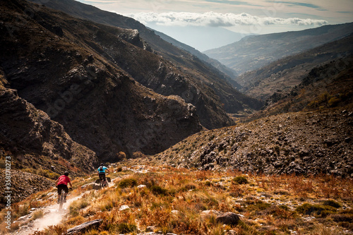 SIERRA NEVADA, SPAIN. Two mountain bikers riding down a rough singletrack trail in a barren remote valley with dramatic sky and haze.