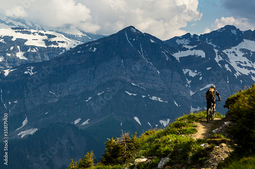 Chatel, Switzerland. A mountain biker riding along a narrow trail on a cliff edge with snowy alpine peaks in the background