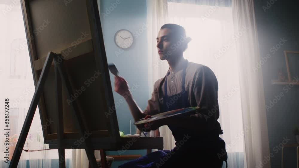 Young artist man is sitting on a stool at home in front of canvas, and working with inspiration, drawing a picture, then looks at camera and smiles, happy to do what he really likes, Slow motion. Young artist man is sitting on a stool at home in front of canvas, and working with inspiration, drawing a picture, then looks at camera and smiles, happy to do what he really likes, Slow motion.
