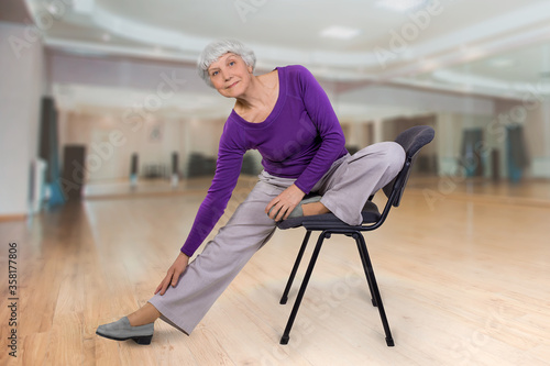 Happy charming beautiful elderly woman is doing exercise on a chair. Exercising gymnastics for health in the fitness room. Sports training.