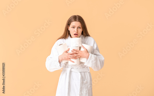 Surprised young woman with many rolls of toilet paper on color background