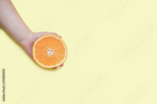 Girl's hand holding orange on yellow background. Festive advertising concept. Vitamin supply and juicy fruits for healthy meal. 