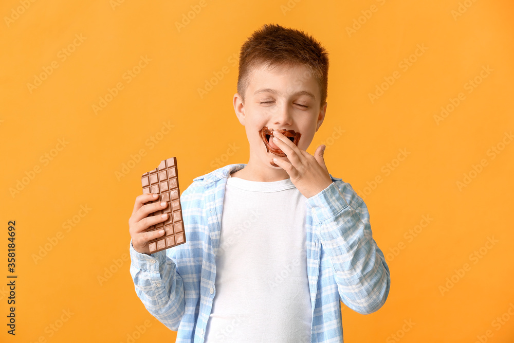 Cute little boy eating chocolate on color background