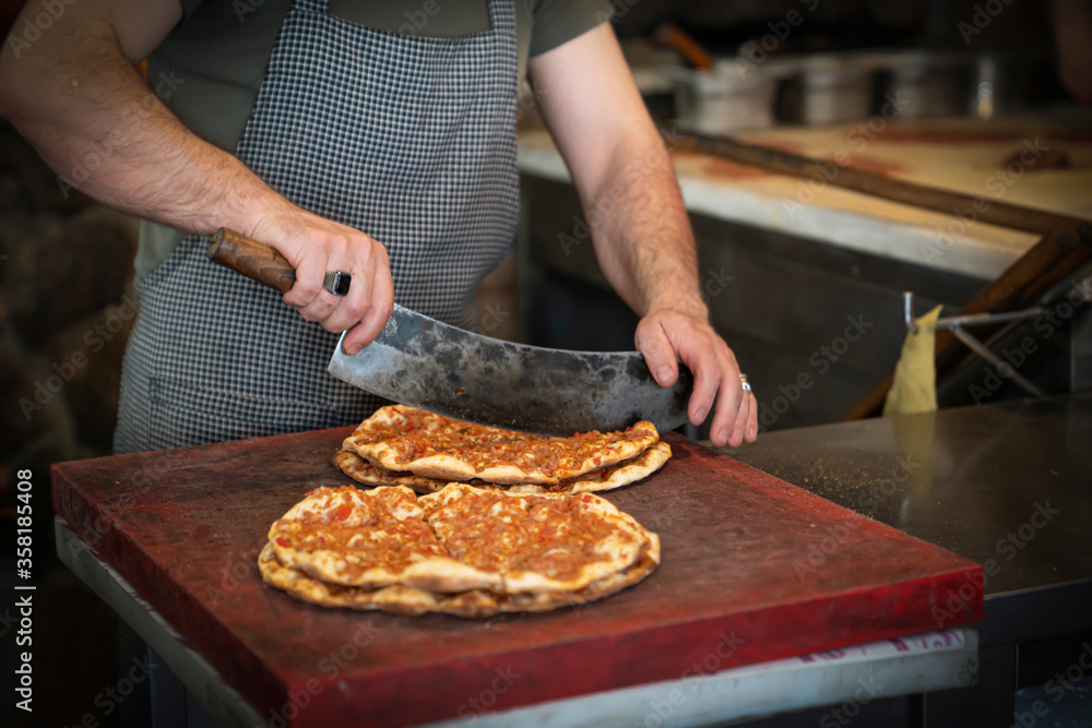 Baked lahmacun is a round, thin piece of dough topped with minced meat (most commonly beef or lamb), minced vegetables and herbs. Man cuts lahmacun.