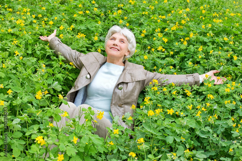 Happy beautiful elderly woman sitting arms outstretched on a glade of yellow flowers in spring