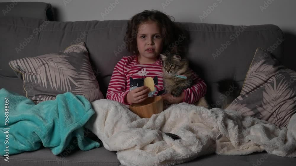 A little girl watches TV at night with a cat and eats chips. A child with a pet on a sofa. Shooting in a dark room. Girl 7 years old European A little girl watches TV at night with a cat and eats chips. A child with a pet on a sofa. Shooting in a dark room. Girl 7 years old European