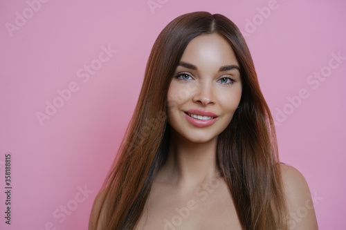 Beautiful long haired woman with pleasant smile, has perfect skin, wears makeup, looks with happy expression, poses against pink background. Female with well cared complexion. Beauty of hair