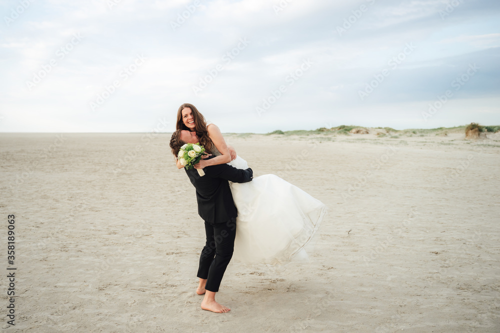 Wedding couple standing and hugging at sandy beach. Groom lifting bride to air. Smiling cheerful bride looking to camera. Wedding day concept. Wedding couple standing and hugging at sandy beach. Groom lifting bride to air. Smiling cheerful bride looking to camera. Wedding day concept.