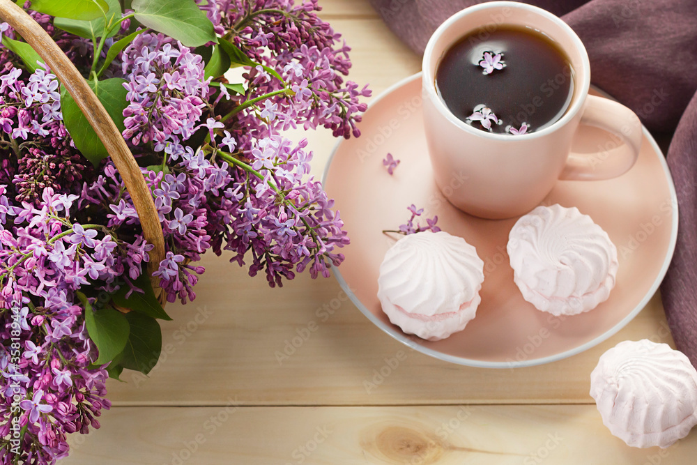 Cup of tea or coffee with sweet pink marshmallows and bouquet of lilac in a basket on wooden background. Breakfast in the morning. Cup of tea or coffee with sweet pink marshmallows and bouquet of lilac in a basket on wooden background. Breakfast in the morning.