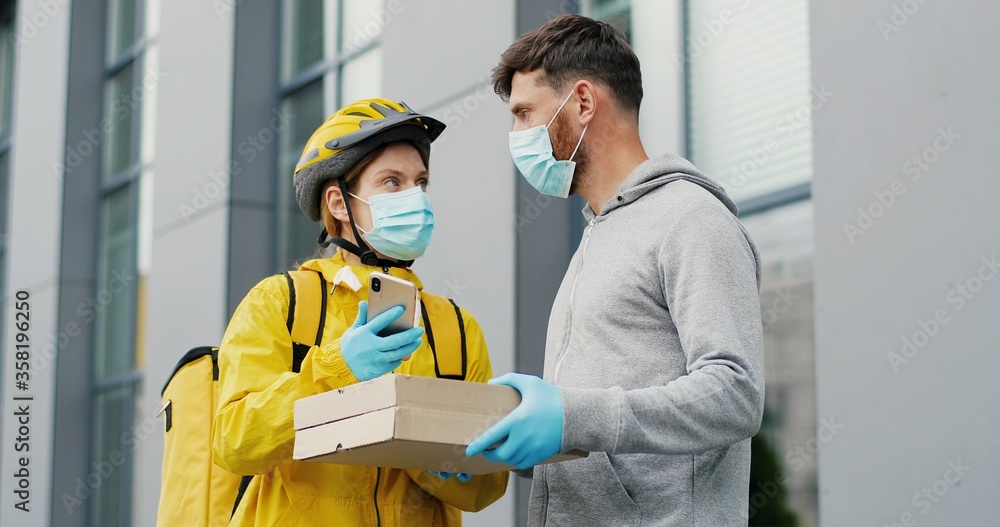 Female courier in medical mask giving carton boxes with pizza to man on street and using smartphone. Food delivery service concept Female courier in medical mask giving carton boxes with pizza to man on street and using smartphone. Food delivery service concept