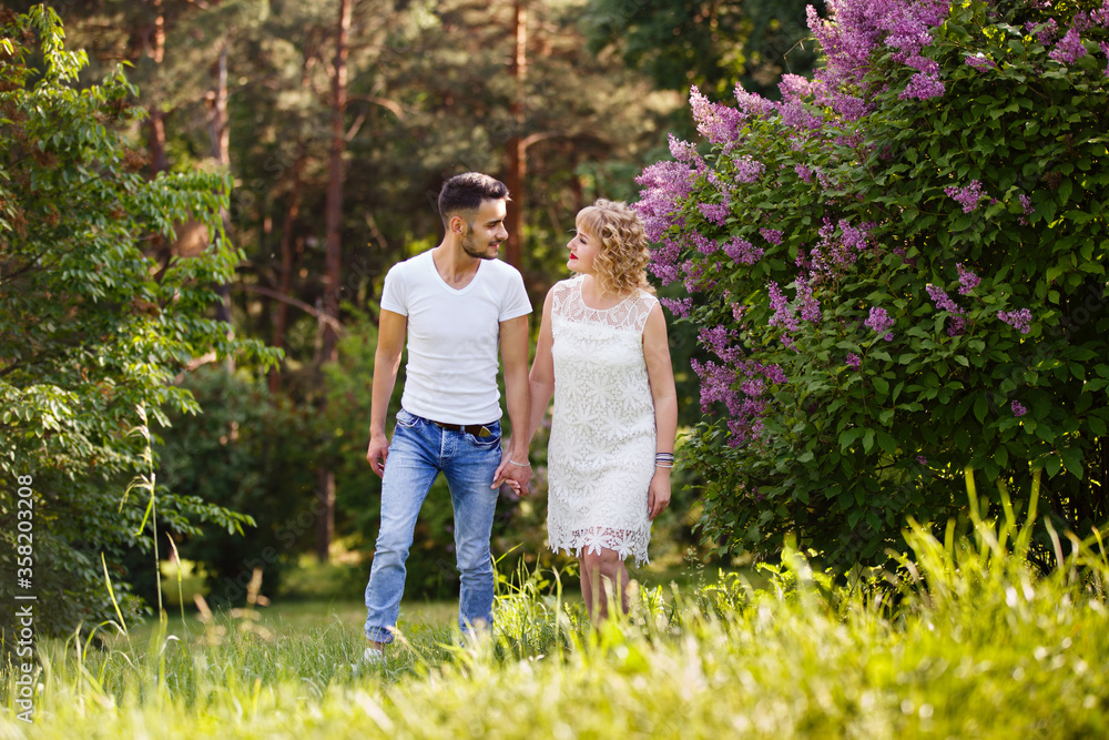 Fototapeta premium Young man and woman couple near a blooming lilac bush. Tender holding each other. Spring lovestory. Blonde-haired girl with curled hairs and man weared in casual. Young family