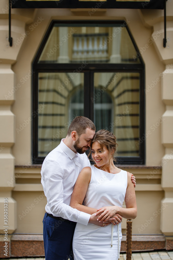 Fototapeta premium Young Bride and Groom couple in summer City. Tender holding each other. Woman and man couple.Romantic date of young happy family on window background. Gender traditional newlyweds street style wedding