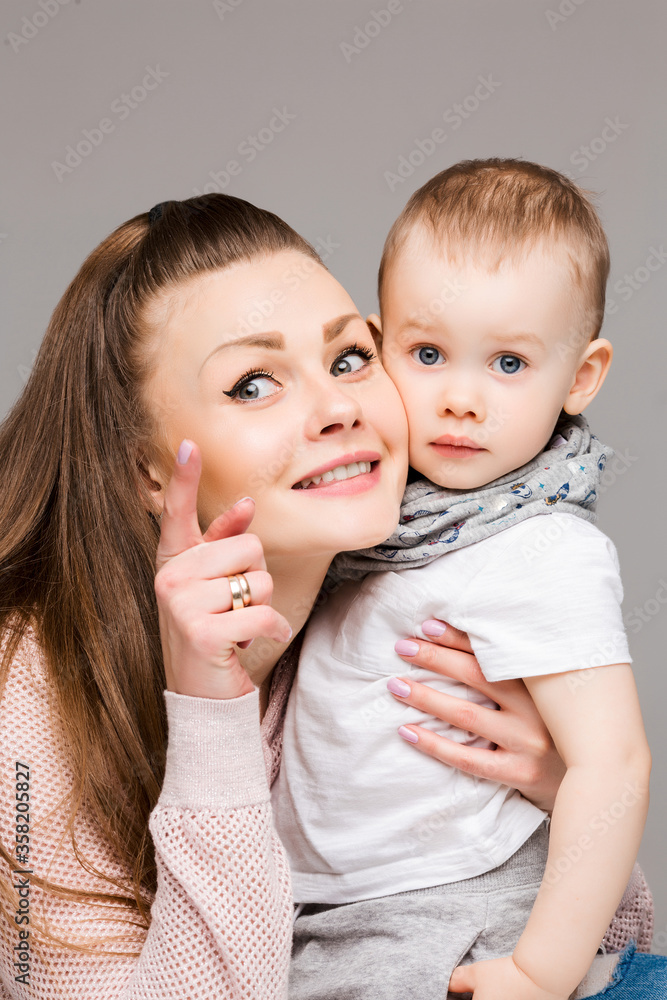Beautiful young mother and little sweet son embracing, woman gesturing and pointing at camera by finger. Smiling stylish family of mom and kid with blue eyes, posing at gray studio. Gray background. Beautiful young mother and little sweet son embracing, woman gesturing and pointing at camera by finger. Smiling stylish family of mom and kid with blue eyes, posing at gray studio. Gray background.