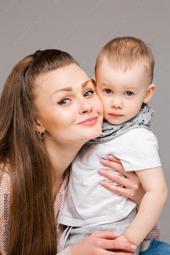 Beautiful young mother and little sweet son embracing, woman gesturing and pointing at camera by finger. Smiling stylish family of mom and kid with blue eyes, posing at gray studio. Gray background. Beautiful young mother and little sweet son embracing, woman gesturing and pointing at camera by finger. Smiling stylish family of mom and kid with blue eyes, posing at gray studio. Gray background.