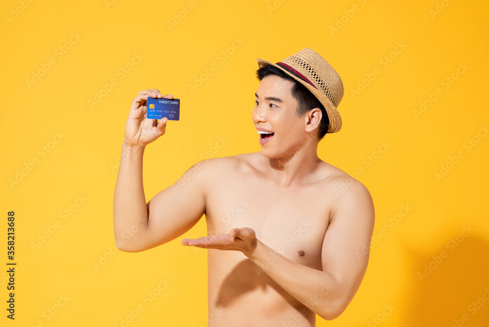 Portrait of a smiling shirtless young Asian tourist man showing credit card in isolated studio yellow background Portrait of a smiling shirtless young Asian tourist man showing credit card in isolated studio yellow background