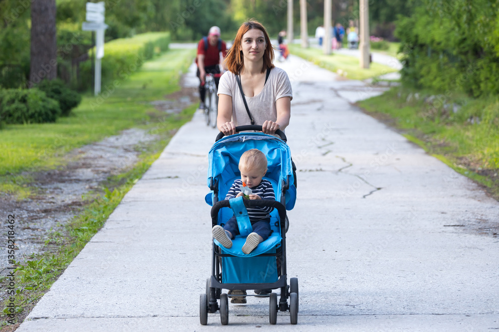 A young mother walks with a stroller in which the one-year-old son cheerfully smiles and eats mash on a warm summer day in a city park A young mother walks with a stroller in which the one-year-old son cheerfully smiles and eats mash on a warm summer day in a city park