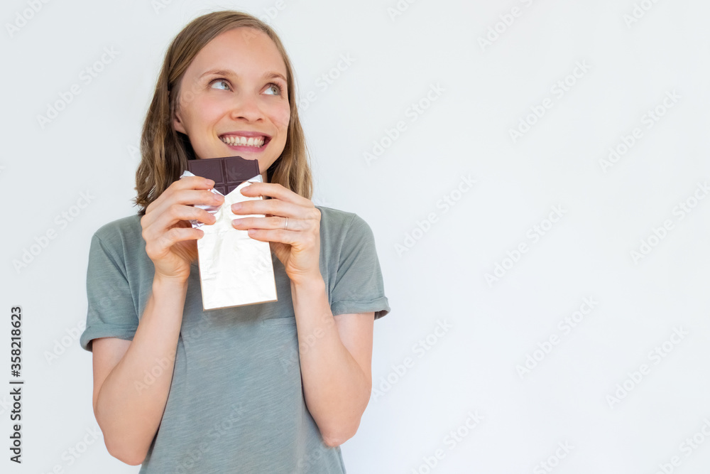 Fascinated young woman holding chocolate bar in gold foil, smiling, enjoying, and looking away. Portrait of girl isolated on white background. Front view. Dessert and sweets concept Fascinated young woman holding chocolate bar in gold foil, smiling, enjoying, and looking away. Portrait of girl isolated on white background. Front view. Dessert and sweets concept