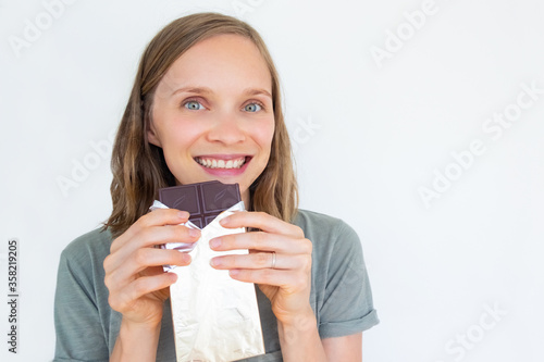 Enthusiastic young woman holding chocolate bar in gold foil, smiling, enjoying, and looking away. Closeup portrait of girl isolated on white background. Front view. Dessert and sweets concept
