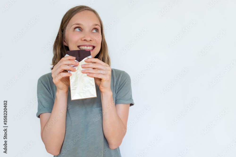 Excited young woman holding chocolate bar in gold foil, smiling, enjoying, and looking away. Portrait of girl isolated on white background. Front view. Dessert and sweets concept Excited young woman holding chocolate bar in gold foil, smiling, enjoying, and looking away. Portrait of girl isolated on white background. Front view. Dessert and sweets concept