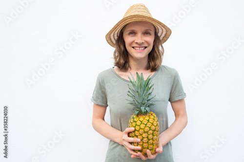 Cheerful young woman in straw hat holding whole pineapple fruit, looking at camera, smiling. Front view, person against white background. Summer or harvesting concept