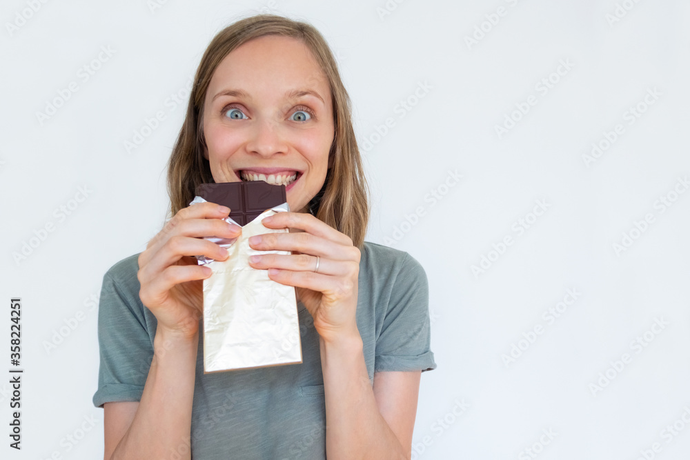 Excited woman eating chocolate bar in gold foil, enjoying, and looking at camera. Closeup portrait of girl isolated on white background. Front view. Dessert and sweets concept Excited woman eating chocolate bar in gold foil, enjoying, and looking at camera. Closeup portrait of girl isolated on white background. Front view. Dessert and sweets concept