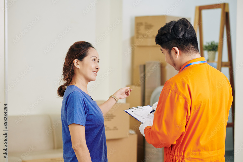 Fototapeta premium Horizontal medium shot of mature Asian woman asking questions to delivery service worker while he is making notes
