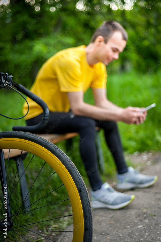 Obraz premium A young man sitting on a bench in a public Park communicates via mobile communication. In the background with his bike. Among trees and vegetation.