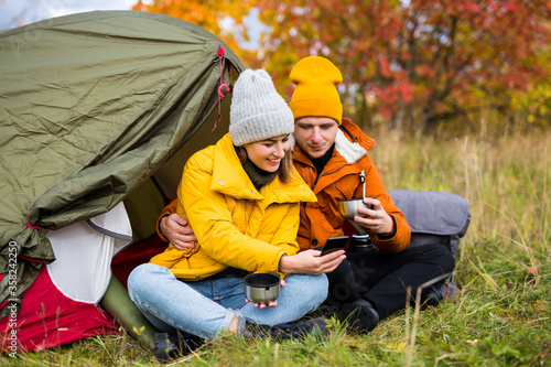 travel, trekking and hiking concept - cute couple sitting near green tent with smart phone and tea in autumn forest