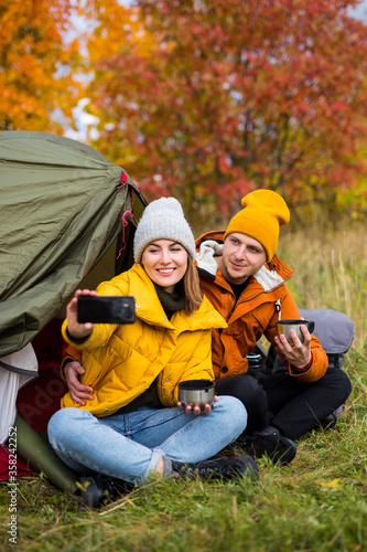 travel, trekking and hiking concept - cute couple sitting near green tent, drinking tea and taking selfie photo in autumn forest