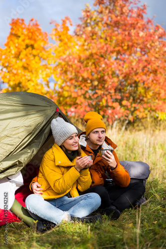 travel, trekking and hiking concept - happy hikers sitting near green tent and drinking tea in autumn forest