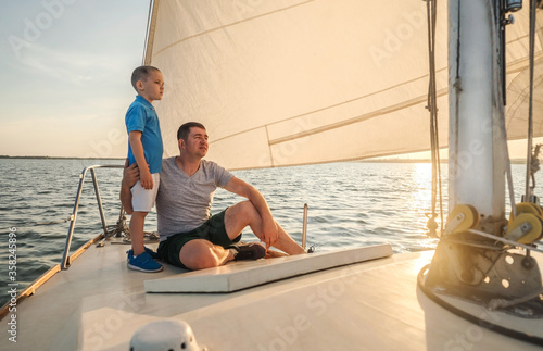 Happy traveler father and son enjoying sunset from deck of sailing boat moving in sea at evening time. Bonding Travel, Summer, Holidays, Journey, Trip, Lifestyle, Yachting concept.