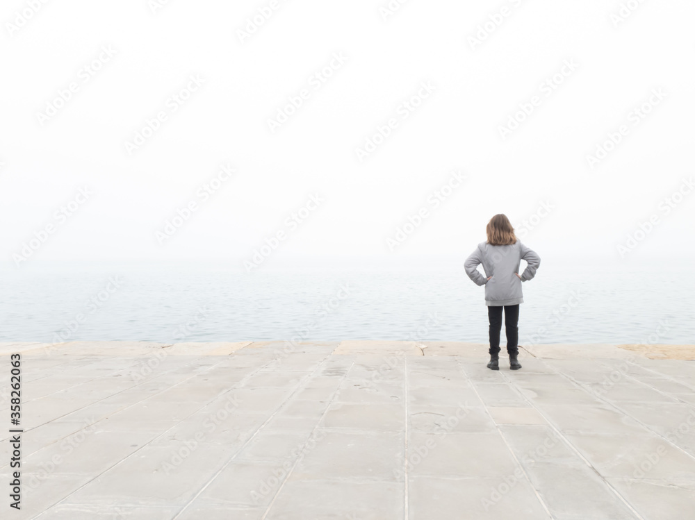Girl child looking at grey distant horizon on a ocean foggy day.