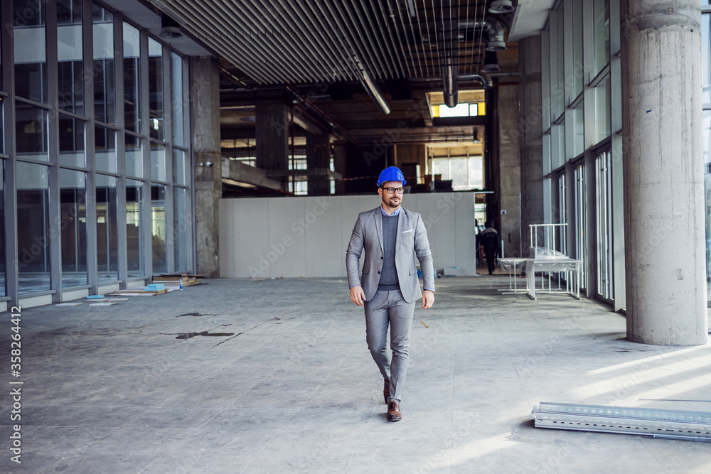 Full length of architect in suit with helmet on head walking around building in construction process.