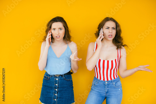 Two pretty confused young girls friends standing isolated over yellow background, talking on mobile phones