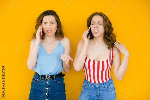 Two pretty confused young girls friends standing isolated over yellow background, talking on mobile phones
