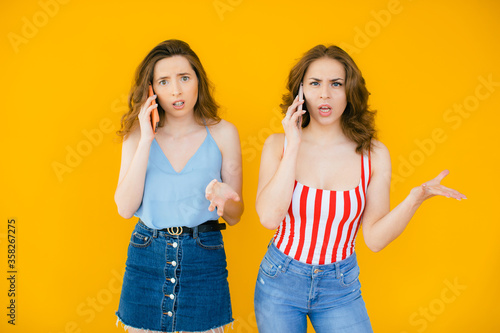 Two pretty confused young girls friends standing isolated over yellow background, talking on mobile phones