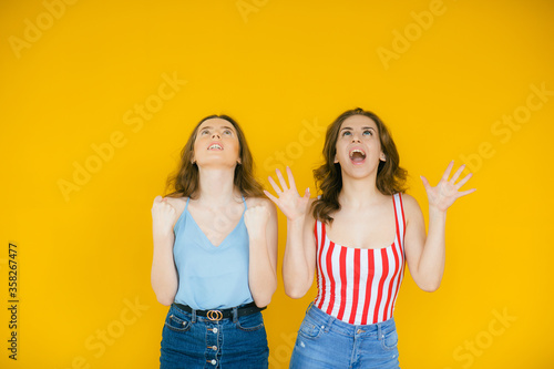 Two excited young girls dressed in summer clothes whispering secrets isolated over yellow background