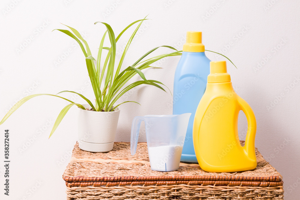 washing powder in a measuring cup, a plant in a pot, washing gel and fabric softener in bright plastic bottles of yellow and blue on a wicker brush, washing colored clothes concept washing powder in a measuring cup, a plant in a pot, washing gel and fabric softener in bright plastic bottles of yellow and blue on a wicker brush, washing colored clothes concept