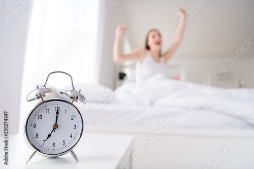 Photo of charming young lady bed white sheets blanket clothes metal alarm clock ringing seven o'clock early morning yawning lazy worker yawning stretching bedroom indoors