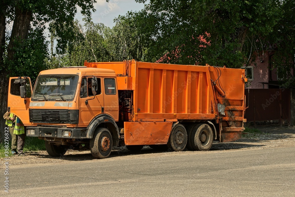 one large orange truck a garbage collector stands on an asphalt road in the street