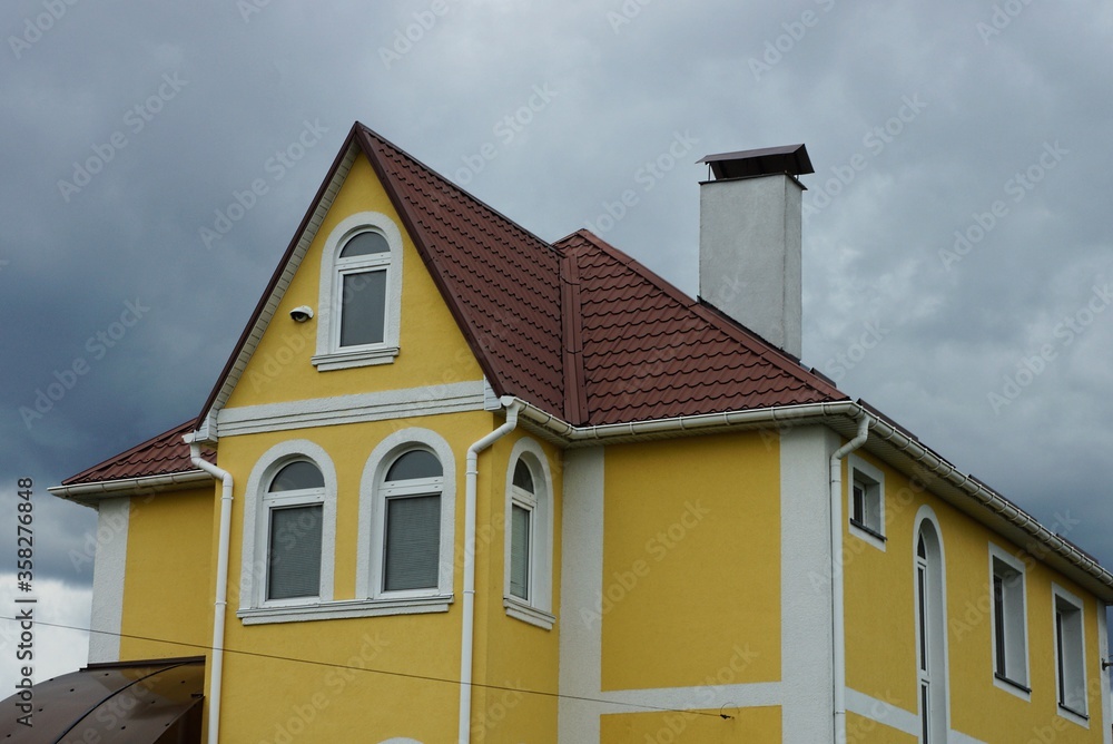 attic of a large yellow private house with white windows under a brown tiled roof against a background of gray clouds