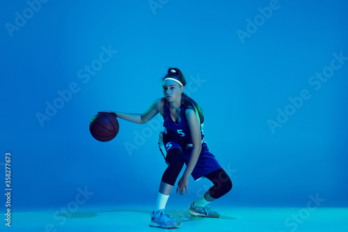 Power of youth. Young caucasian female basketball player training, prcticing with ball isolated on blue background in neon light. Concept of sport, movement, energy and dynamic, healthy lifestyle.