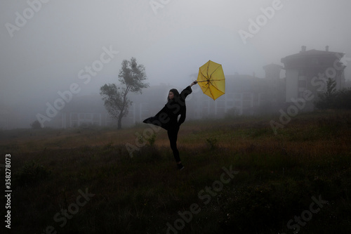 Lonely woman in black jacket walks with yellow umbrella through the fog in forest.