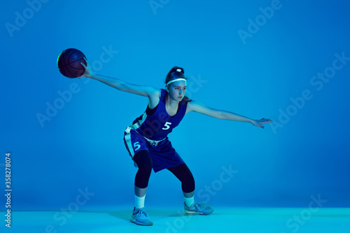 Power of youth. Young caucasian female basketball player training, prcticing with ball isolated on blue background in neon light. Concept of sport, movement, energy and dynamic, healthy lifestyle.