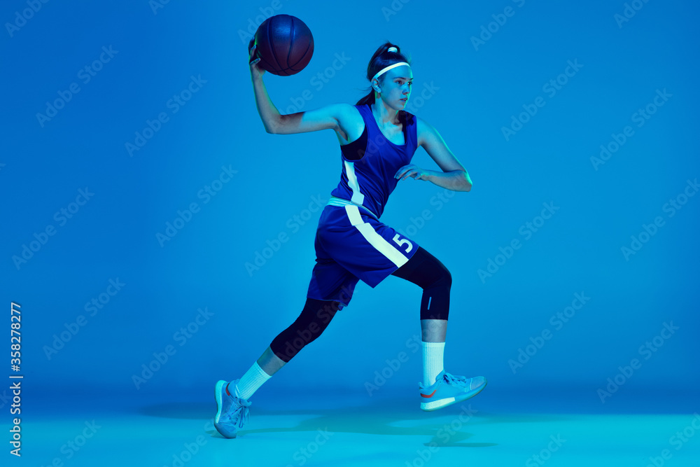 Desire to win. Young caucasian female basketball player training, prcticing with ball isolated on blue background in neon light. Concept of sport, movement, energy and dynamic, healthy lifestyle.