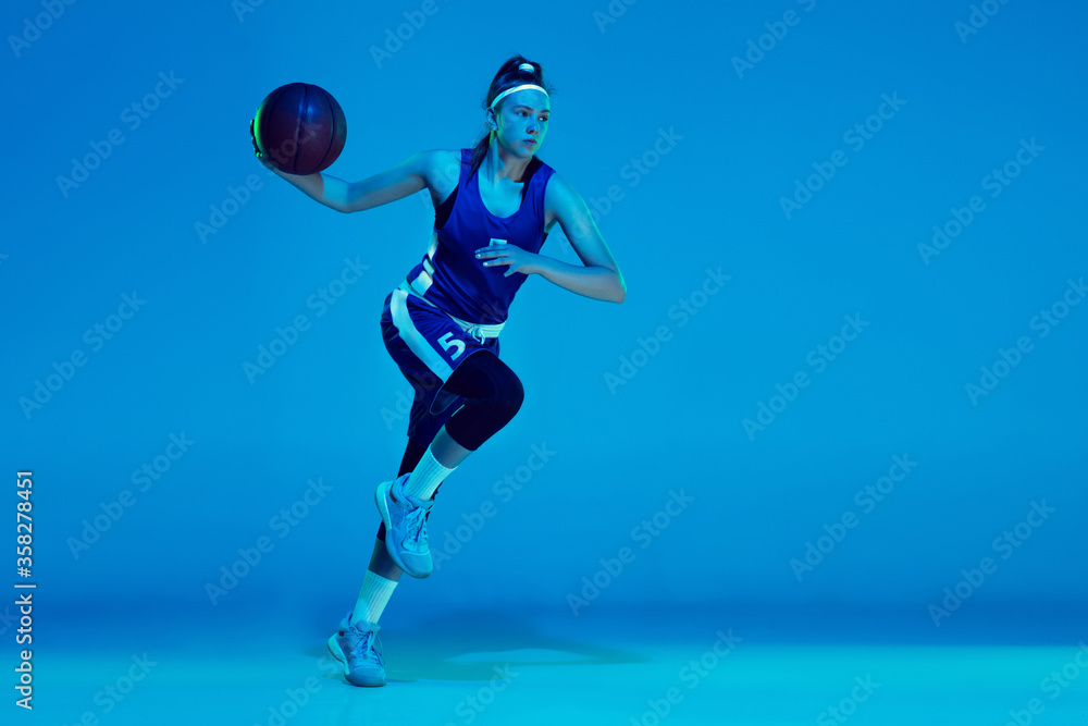 Desire to win. Young caucasian female basketball player training, prcticing with ball isolated on blue background in neon light. Concept of sport, movement, energy and dynamic, healthy lifestyle.