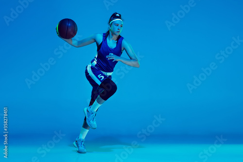 Desire to win. Young caucasian female basketball player training, prcticing with ball isolated on blue background in neon light. Concept of sport, movement, energy and dynamic, healthy lifestyle.