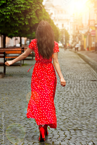 Happy girl walks down the street with cobblestones. Woman in a red dress with white spots. Dresses and hair are blown away by the wind. Girl on a background of blurred city street. © Ivan