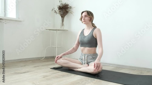 young fit pretty girl in sportswear sits in lotus pose with crossed legs, sets timer on smartphone, closes eyes, starts meditating in bright room at home. Healthy lifestyle, stress relief, meditation