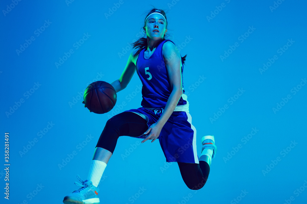 Unstoppable. Young caucasian female basketball player training, prcticing with ball isolated on blue background in neon light. Concept of sport, movement, energy and dynamic, healthy lifestyle.